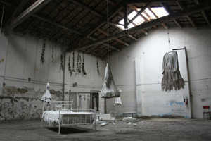 Foto di interno di fabbrica dismessa con all'interno un letto, vestiti appesi e altri oggetti di colore bianco
