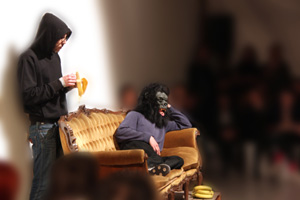 Uomo con la maschera di gorilla seduto su divano e altro uomo dietro che mangia una banana 
